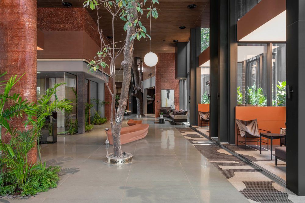 Elegant open lobby featuring indoor trees, polished concrete floors, pendant lights, and orange lounge chairs.