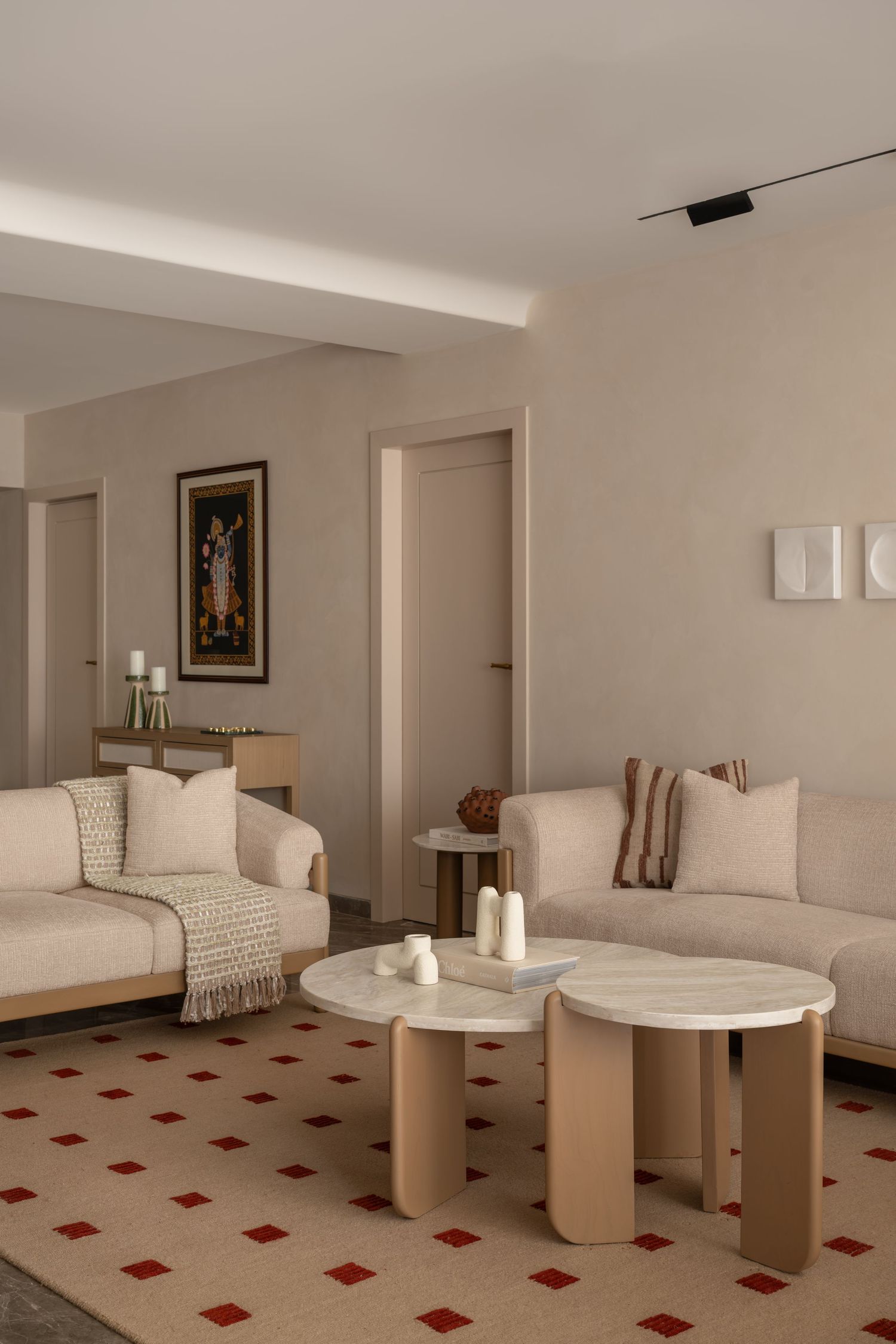 Neutral living room with beige sofas, round marble coffee tables, red diamond-patterned rug, and framed artwork.