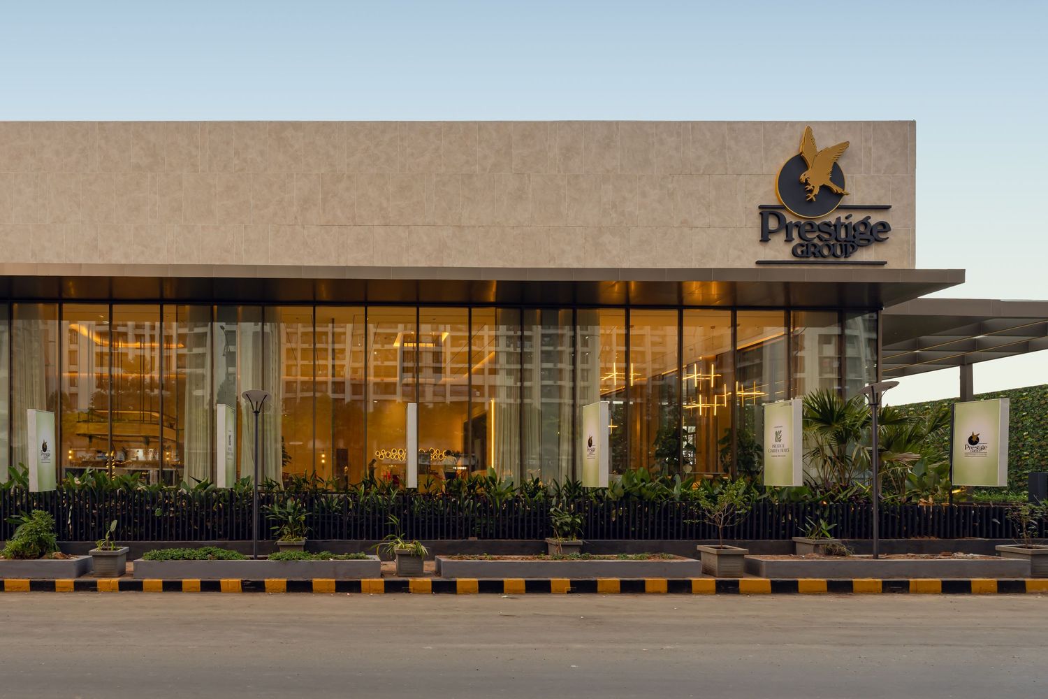 Prestige Group modern showroom exterior with glass facade, landscaped greenery, and illuminated interior at dusk.