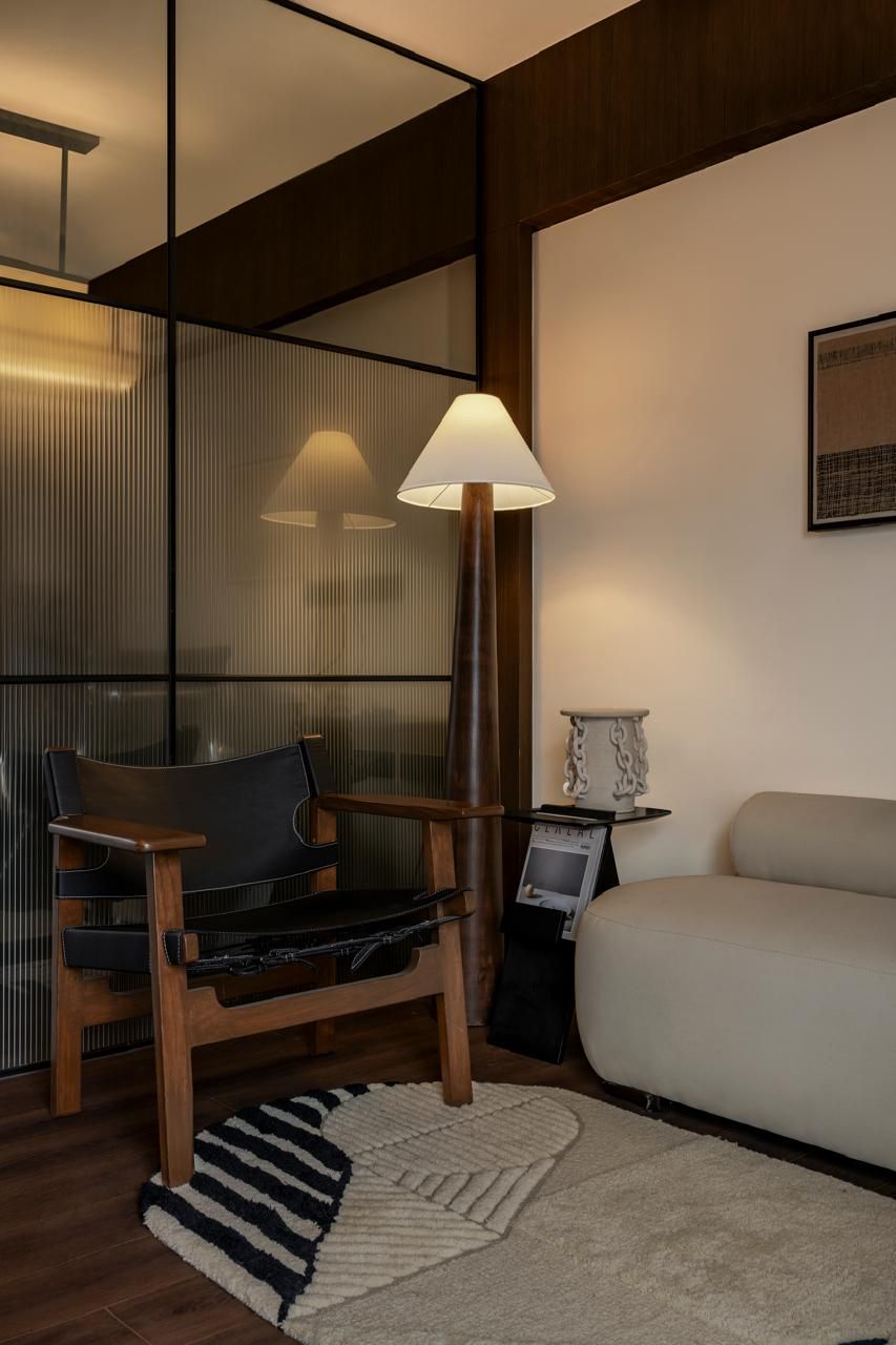 Modern living room corner with leather chair, floor lamp, glass partition, and neutral sofa on textured rug.