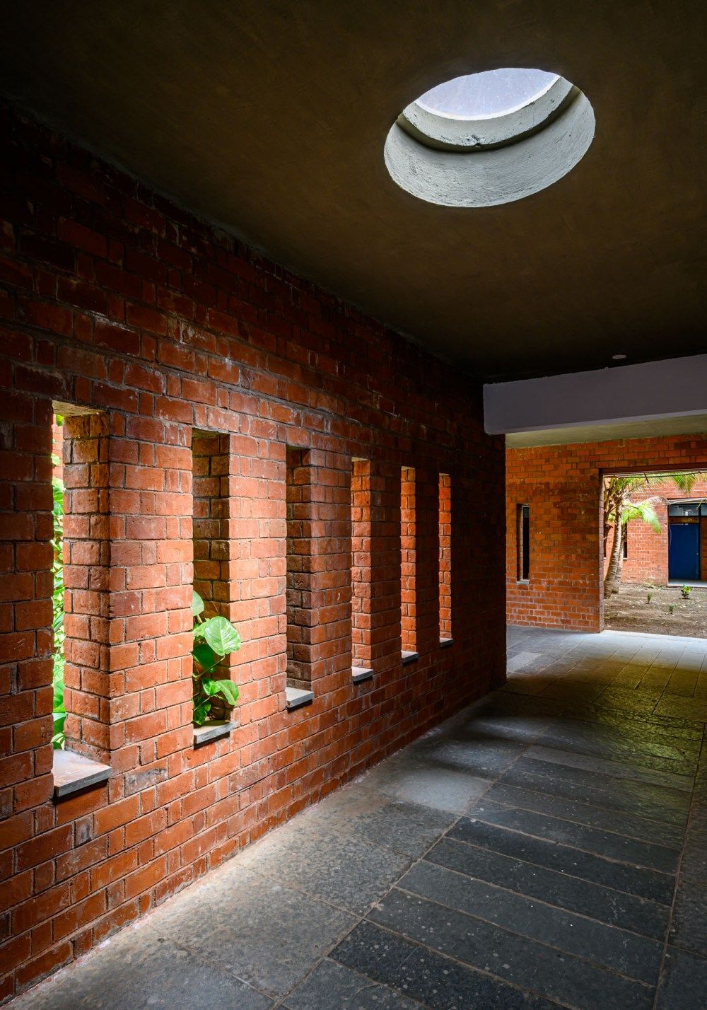 Red brick corridor with rectangular window niches, dark ceiling with circular skylight, and stone tile flooring in modern architecture.