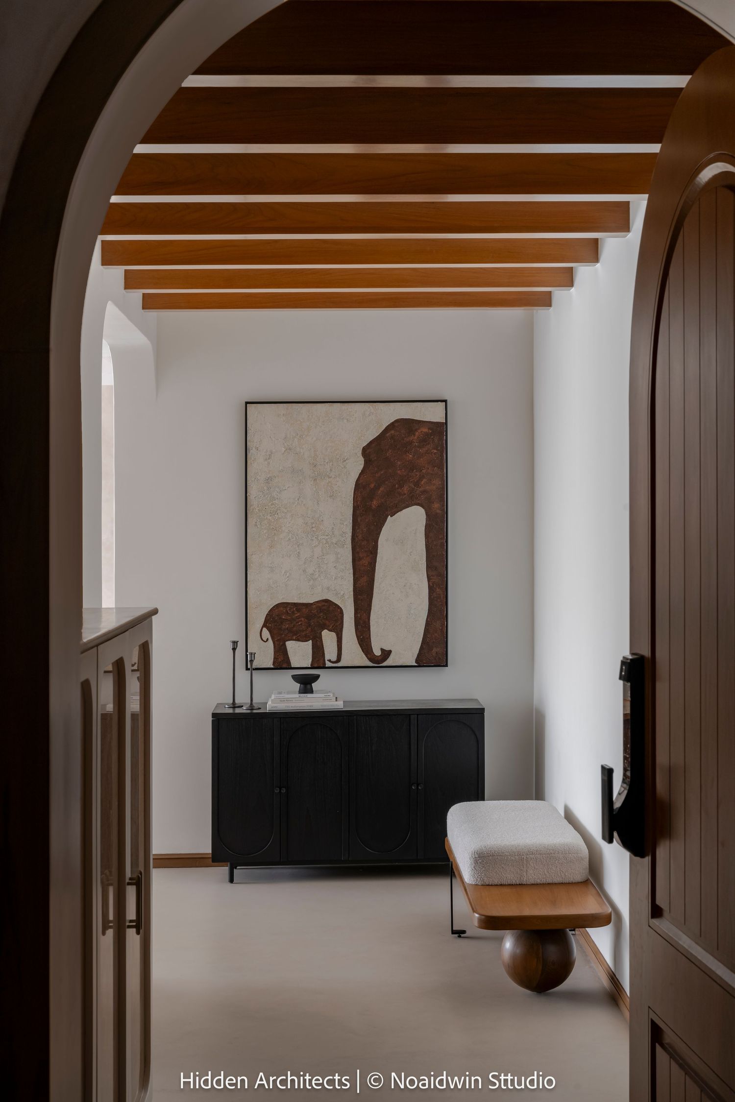 Elegant entryway viewed through arched door with exposed wooden beams, elephant artwork, black console, and upholstered bench.