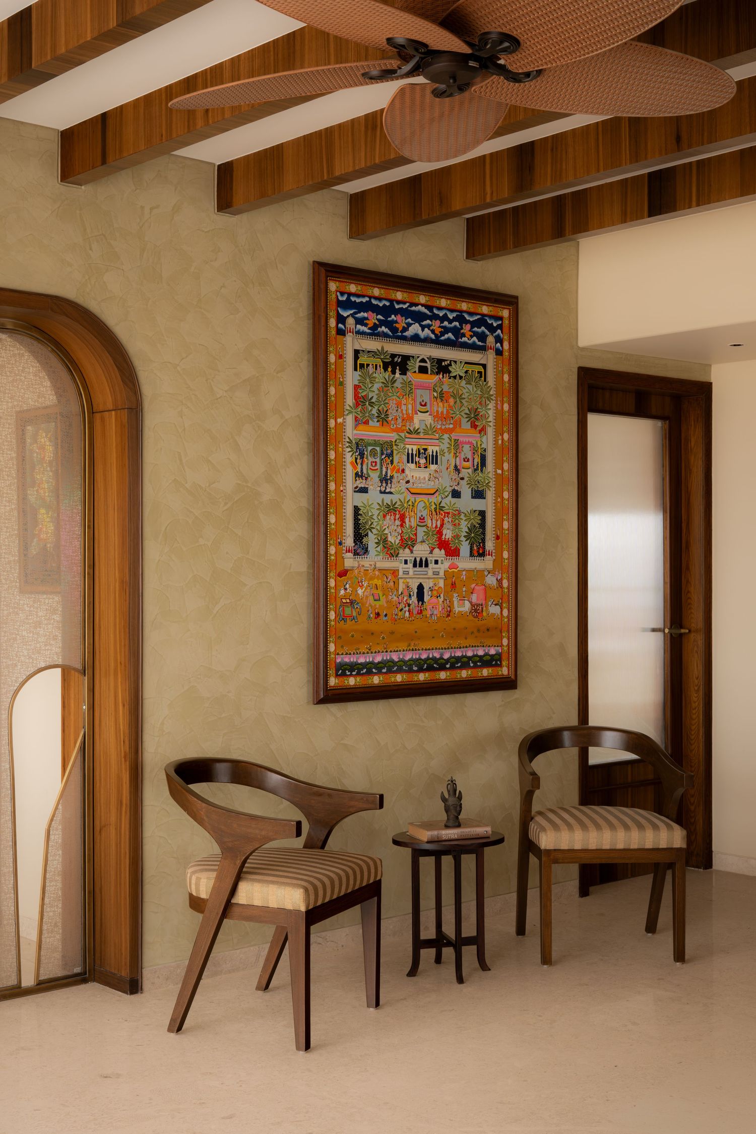 Indian-style hallway with wooden ceiling beams, traditional Pichwai painting, arched door, and carved wooden chairs.