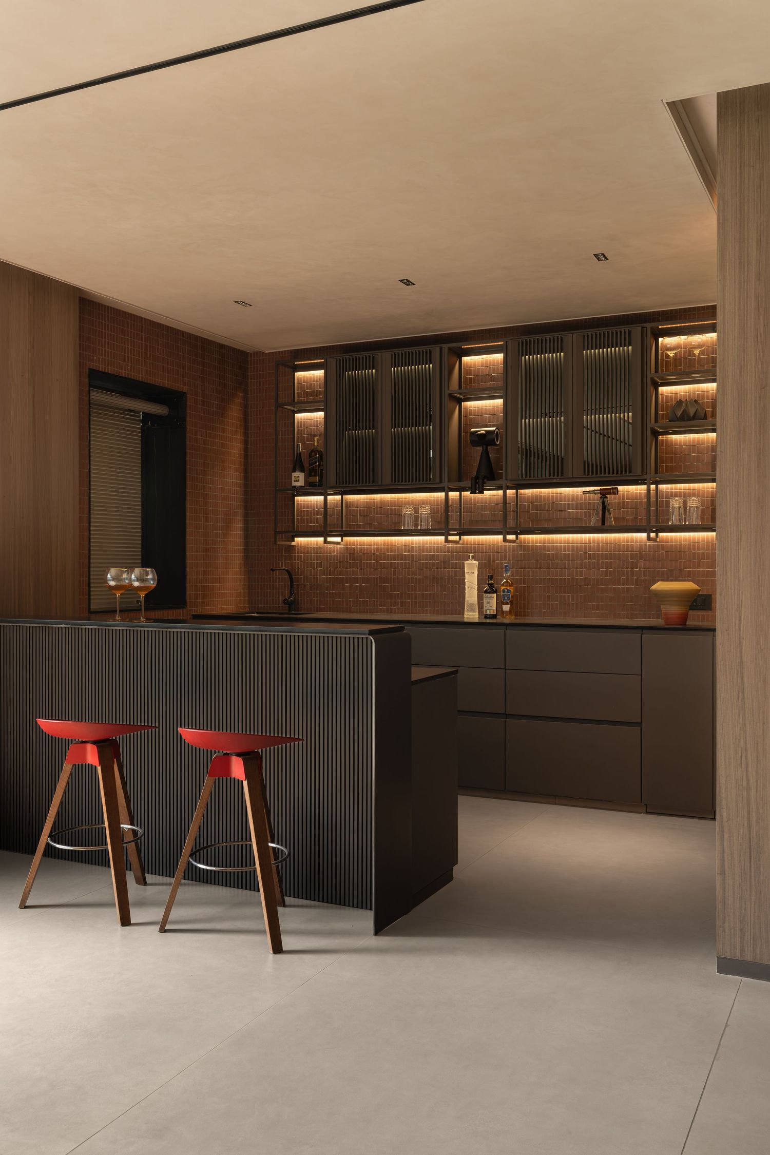 Modern dark kitchen with black cabinetry, glass-front upper cabinets, warm LED lighting, and two red bar stools.