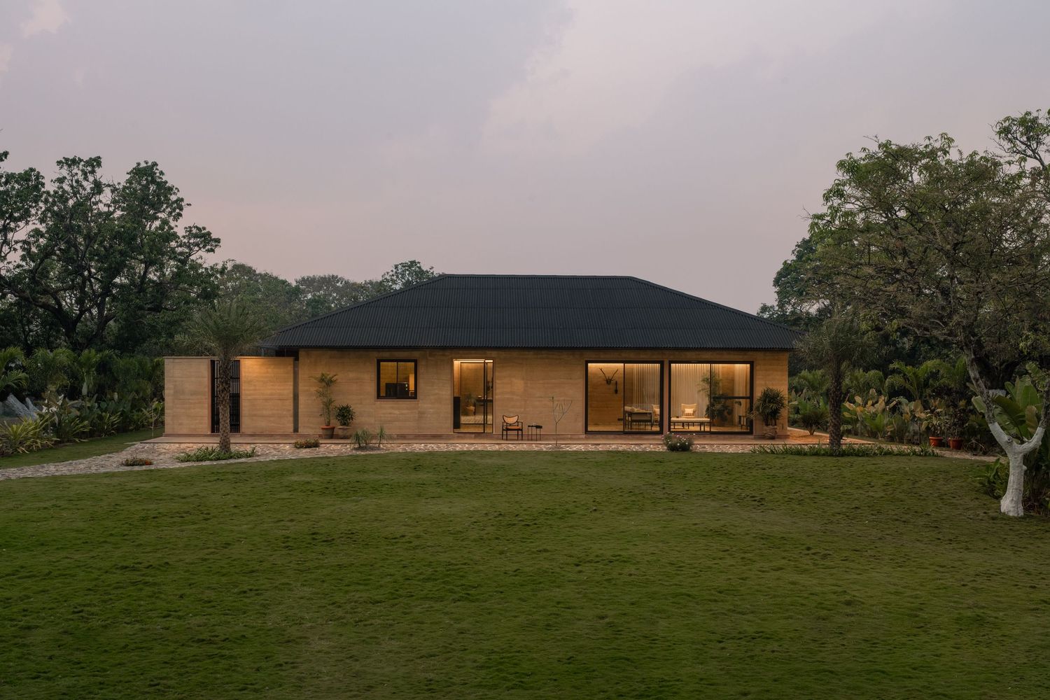 Single-story modern farmhouse with dark roof glowing warmly at twilight on green lawn.