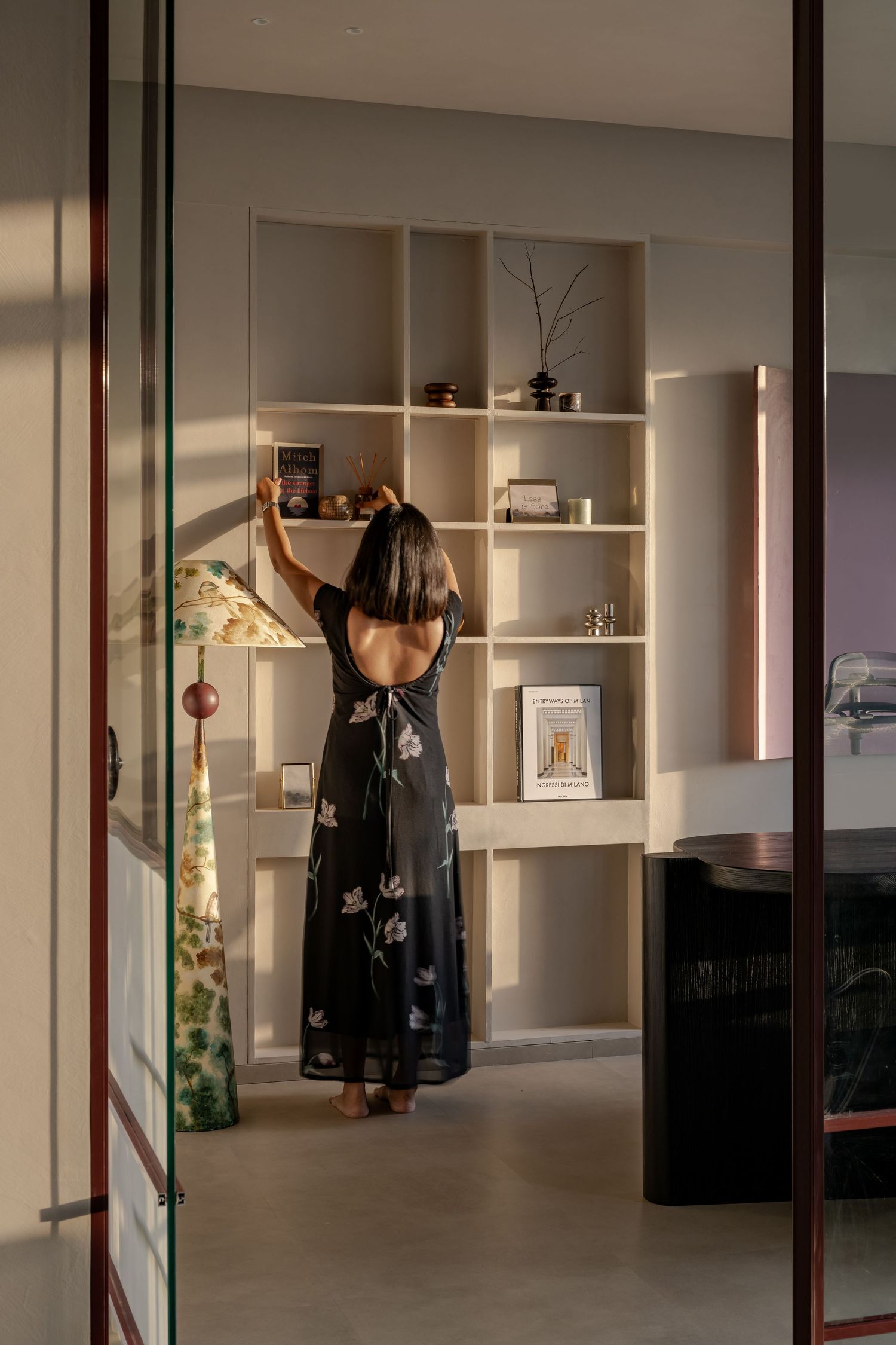 Woman in floral dress arranging items on cream built-in shelving unit in a warmly lit modern interior.