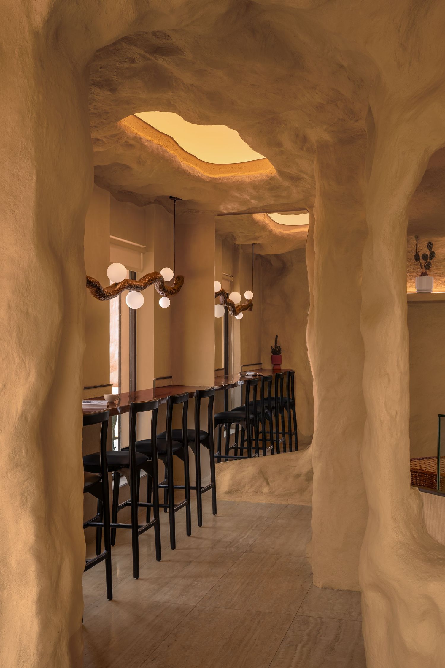 Cave-like restaurant corridor with sculpted sandy plaster walls, arched openings, bar seating, and warm globe pendant lights.