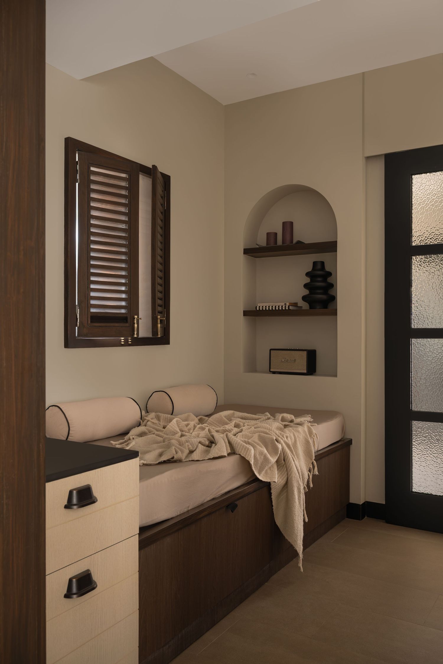 Cozy reading nook with dark wood shutters, built-in arched shelves, and neutral cushioned daybed.
