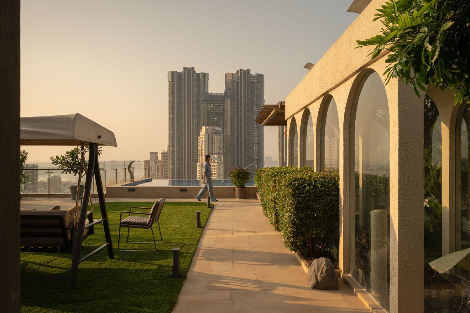Serene rooftop garden walkway with arched colonnade, trimmed hedges, lawn, and high-rise city views at sunset.