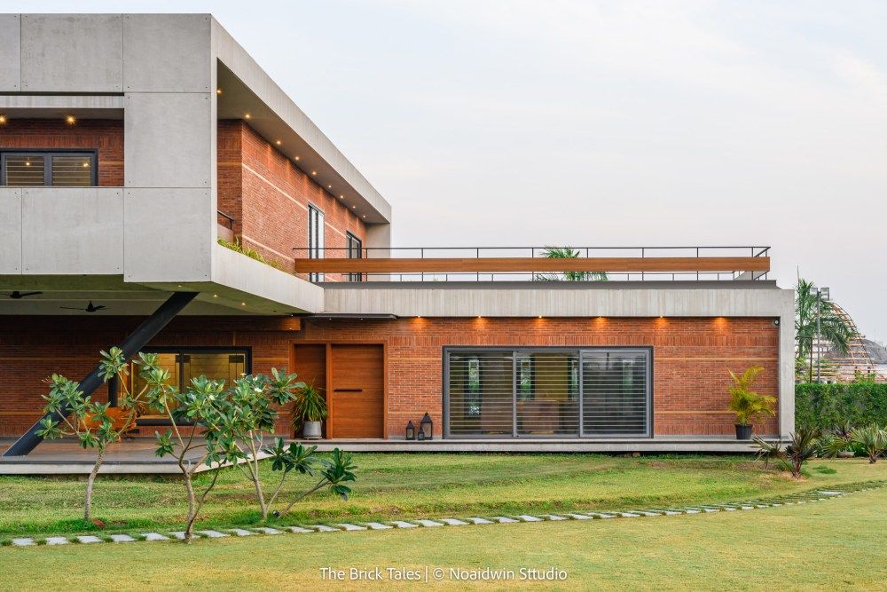 Contemporary brick facade home with large glass sliding doors, manicured lawn, and frangipani trees in India.
