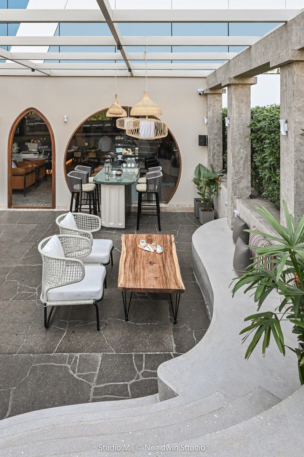 Outdoor restaurant terrace with arched window, wicker pendant lights, rope chairs, live-edge wood coffee table, and stone flooring.