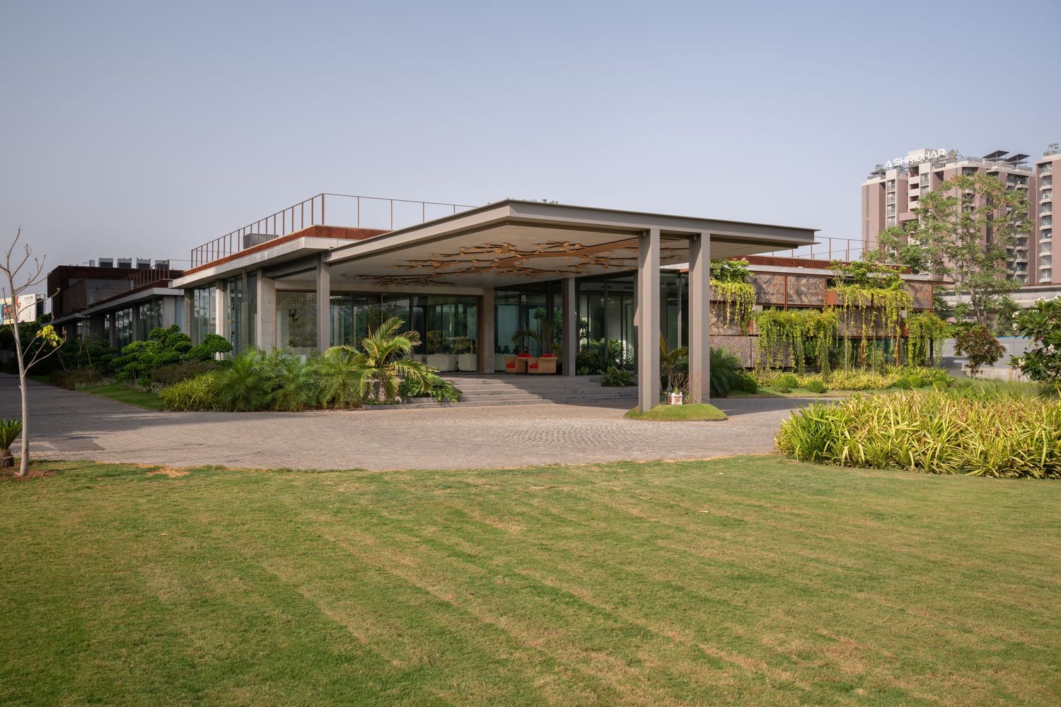 Modern flat-roof building with open pavilion design surrounded by lush green lawn and tropical landscaping.