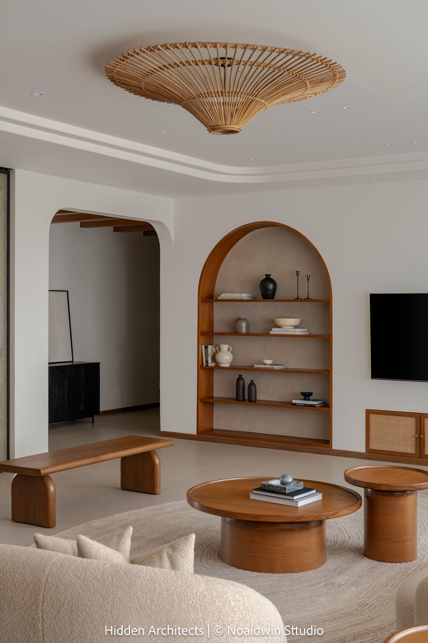 Warm neutral living room with arched wood shelving, rattan ceiling fixture, round walnut coffee tables, and curved beige seating.