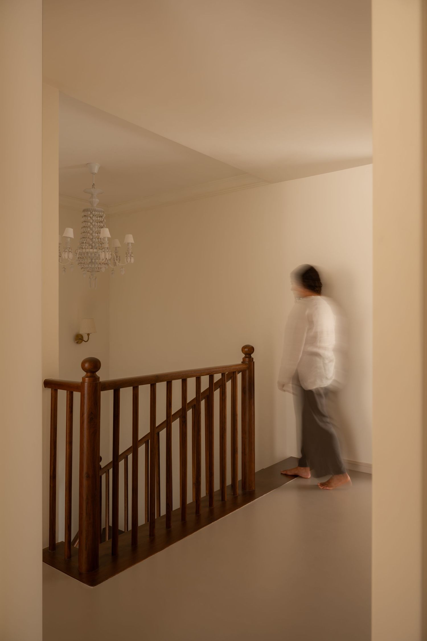 Blurred figure walking past wooden staircase railing beneath a crystal chandelier in a warm neutral hallway.