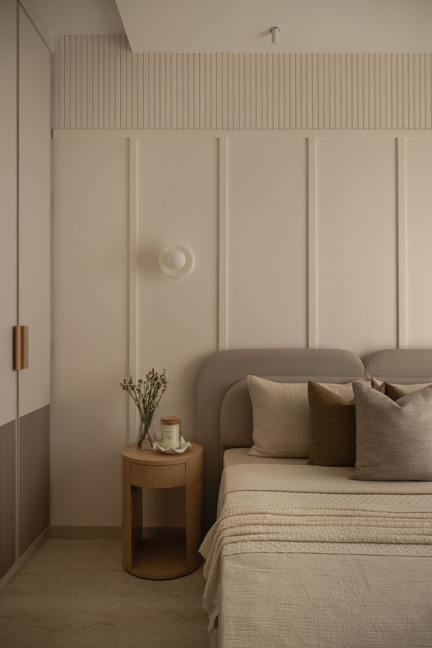 Neutral bedroom with upholstered headboard, round oak nightstand, and decorative wall paneling detail.