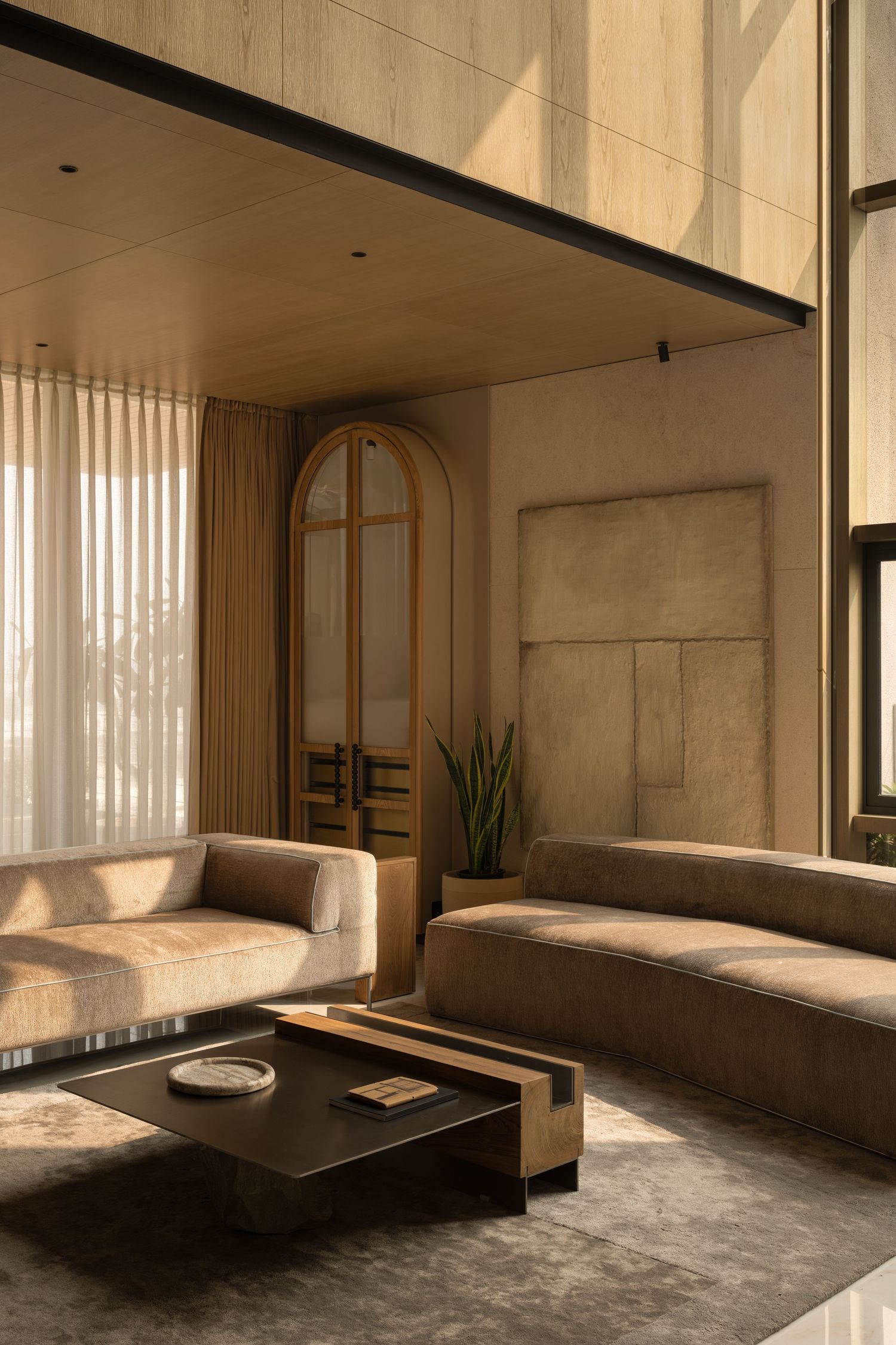 Warm-toned living room with beige sofas, dark coffee table, arched wooden doors, and tall sheer curtains.