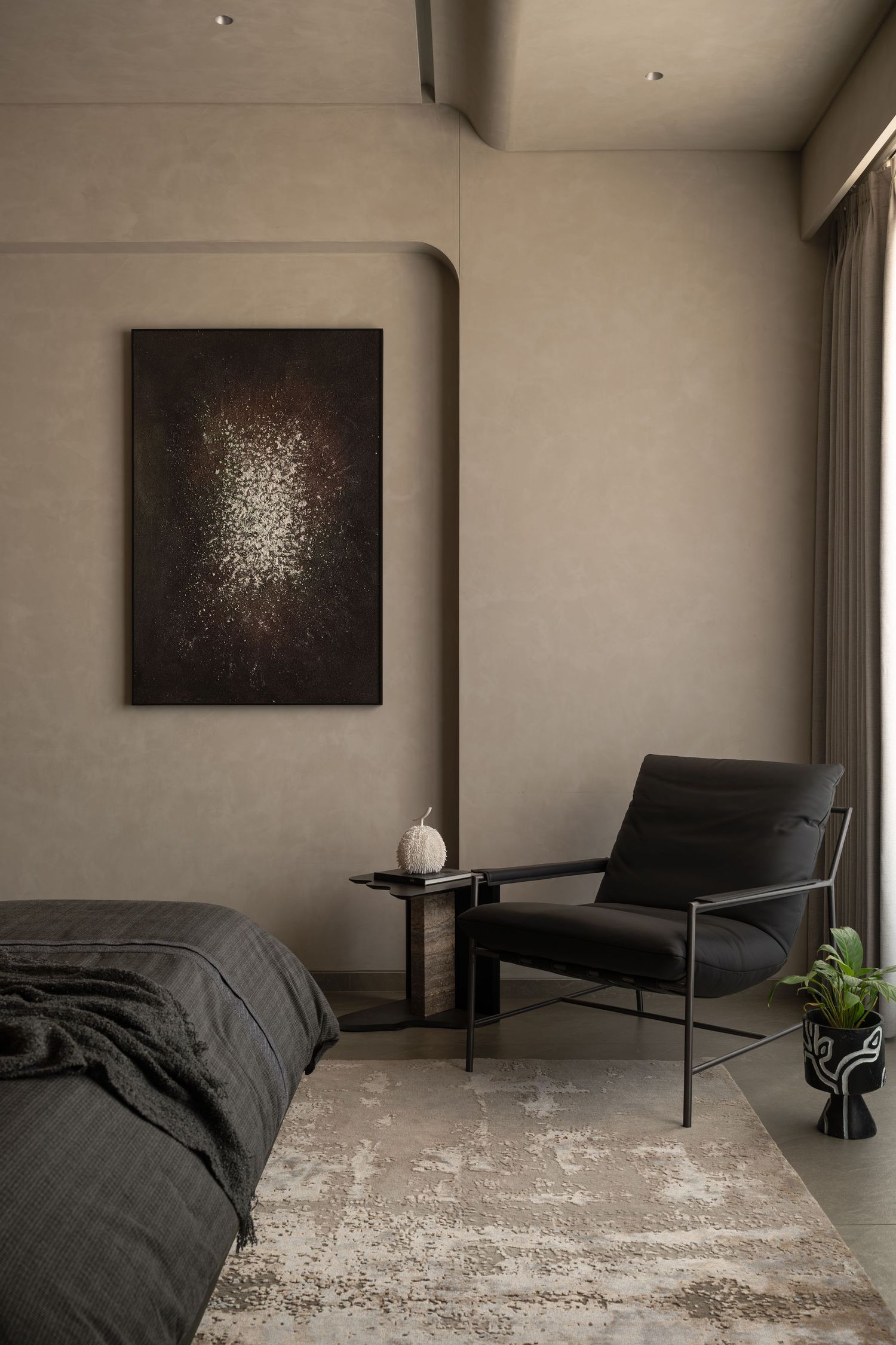 Contemporary bedroom with dark upholstered bed, abstract dark artwork, black metal armchair, and distressed area rug.