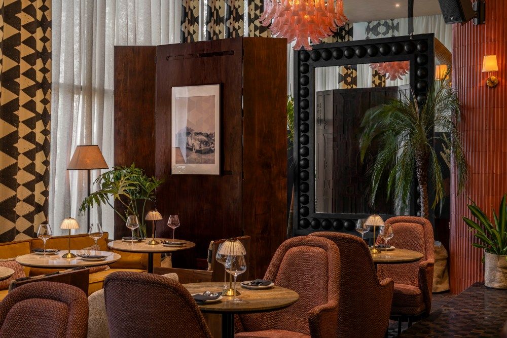 Intimate restaurant setting with velvet chairs, ornate mirror, tropical plant and geometric patterned curtains.