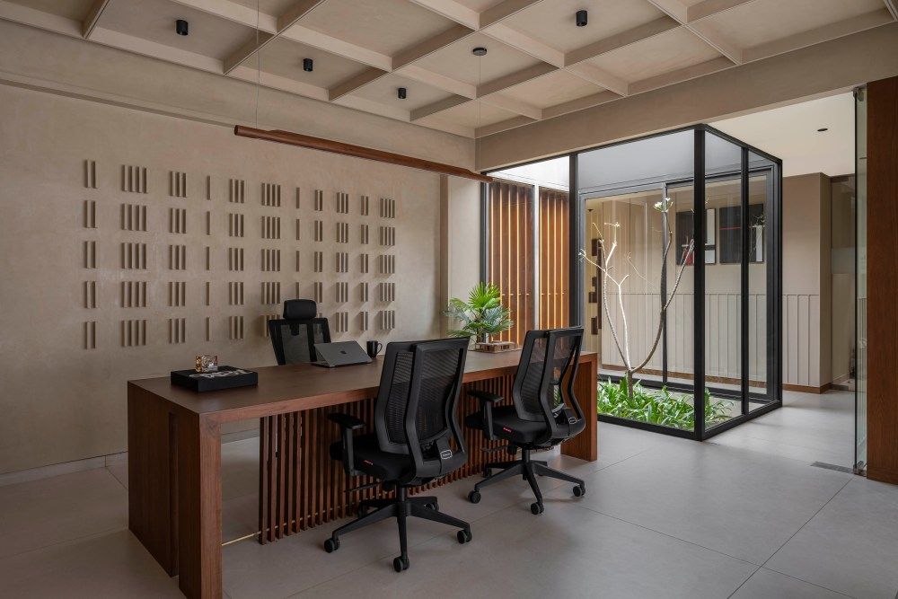 Minimalist office workspace with wooden desk, black ergonomic chairs, textured wall panel, and glass courtyard view.