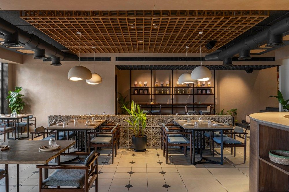 Modern restaurant interior with wooden lattice ceiling, pendant lights, gabion dividers, and dark wood dining chairs on tiled floors.