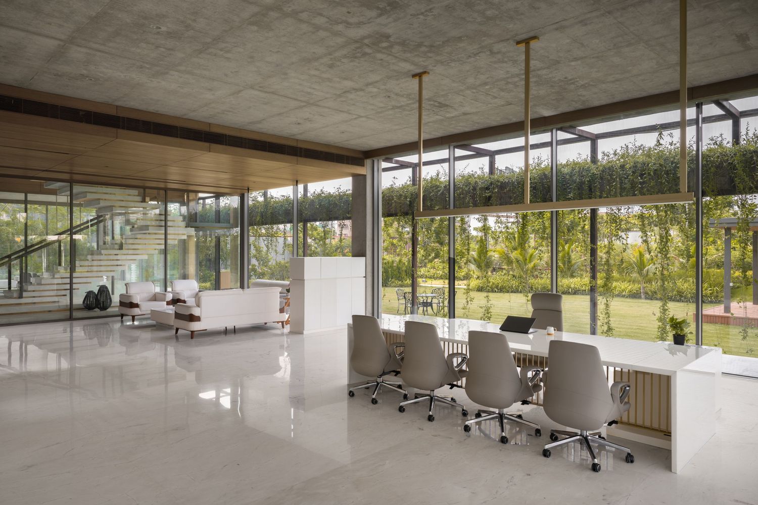 Modern open-plan office with floor-to-ceiling glass walls, conference table, and lush garden views under a concrete ceiling.