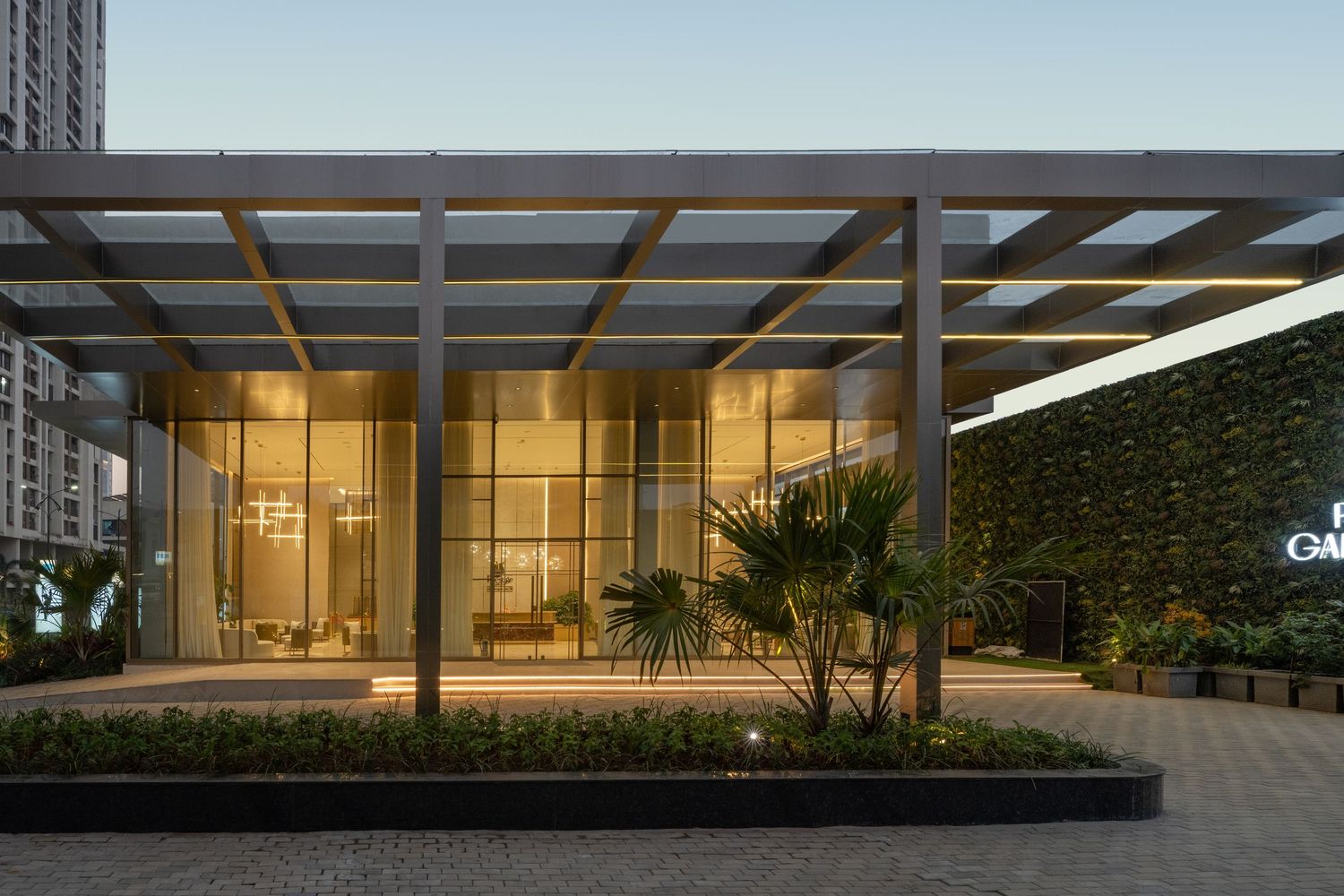 Contemporary glass building entrance with steel pergola, tropical landscaping, and warm interior lighting at dusk.