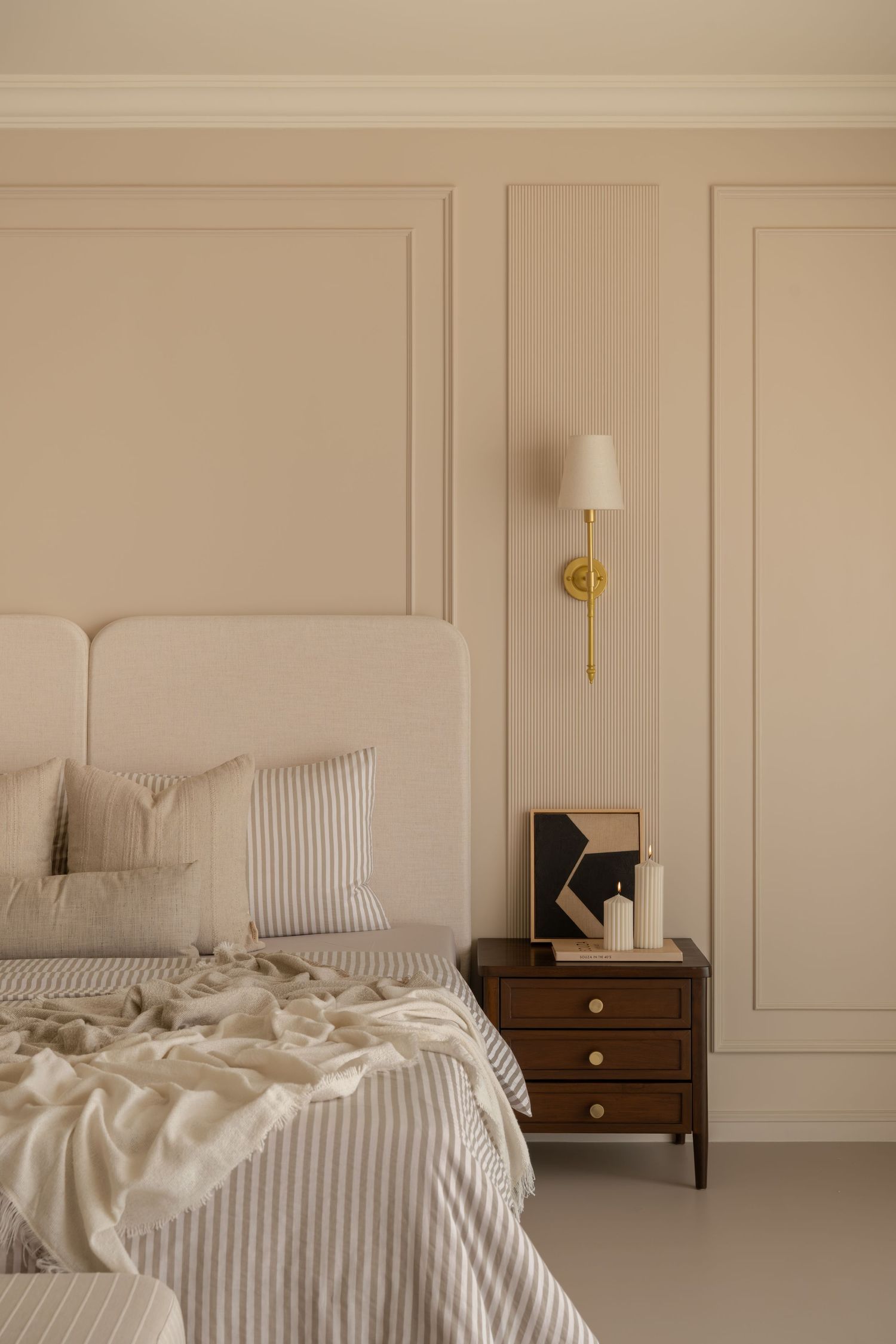 Elegant beige bedroom with upholstered headboard, striped bedding, dark wood nightstand, and gold wall sconce.