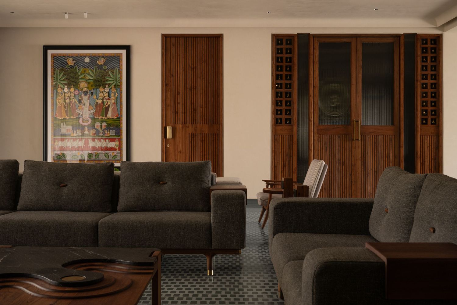 Contemporary living room featuring dark gray sofas, wooden coffee table, traditional Indian wall art, and ornate cabinet.