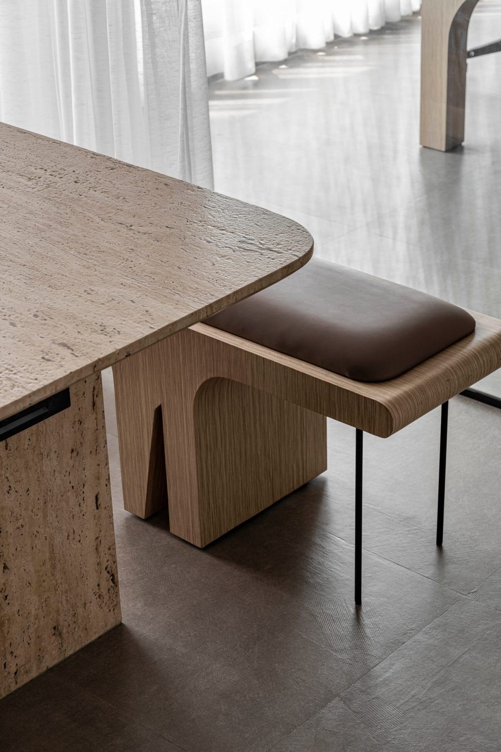 Modern minimalist dining area featuring a travertine table with oak wood base and a brown leather stool on black metal legs.