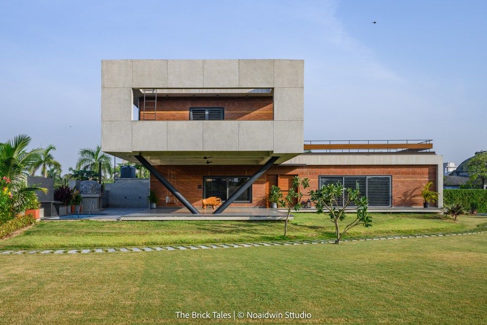 Modern two-story residence featuring concrete upper volume, brick lower level, diagonal steel supports, and green lawn.