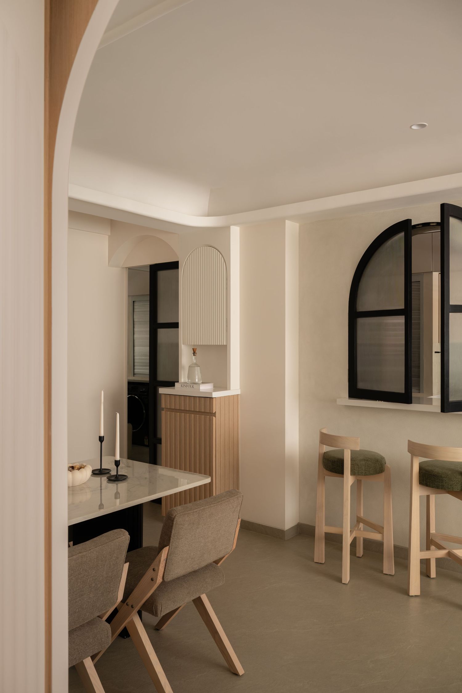 Modern minimalist dining area with arched windows, marble table, wooden chairs, and bar stools in warm neutral tones.