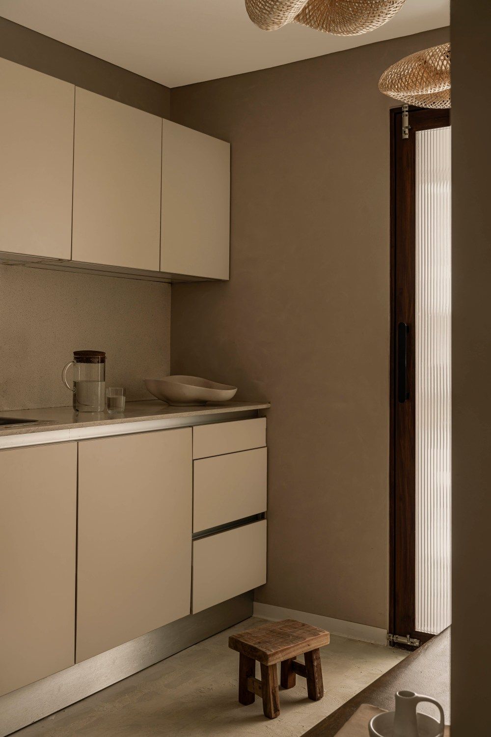 Minimalist kitchen corner with cream cabinetry, stone countertop, woven pendant light, and wooden stool.