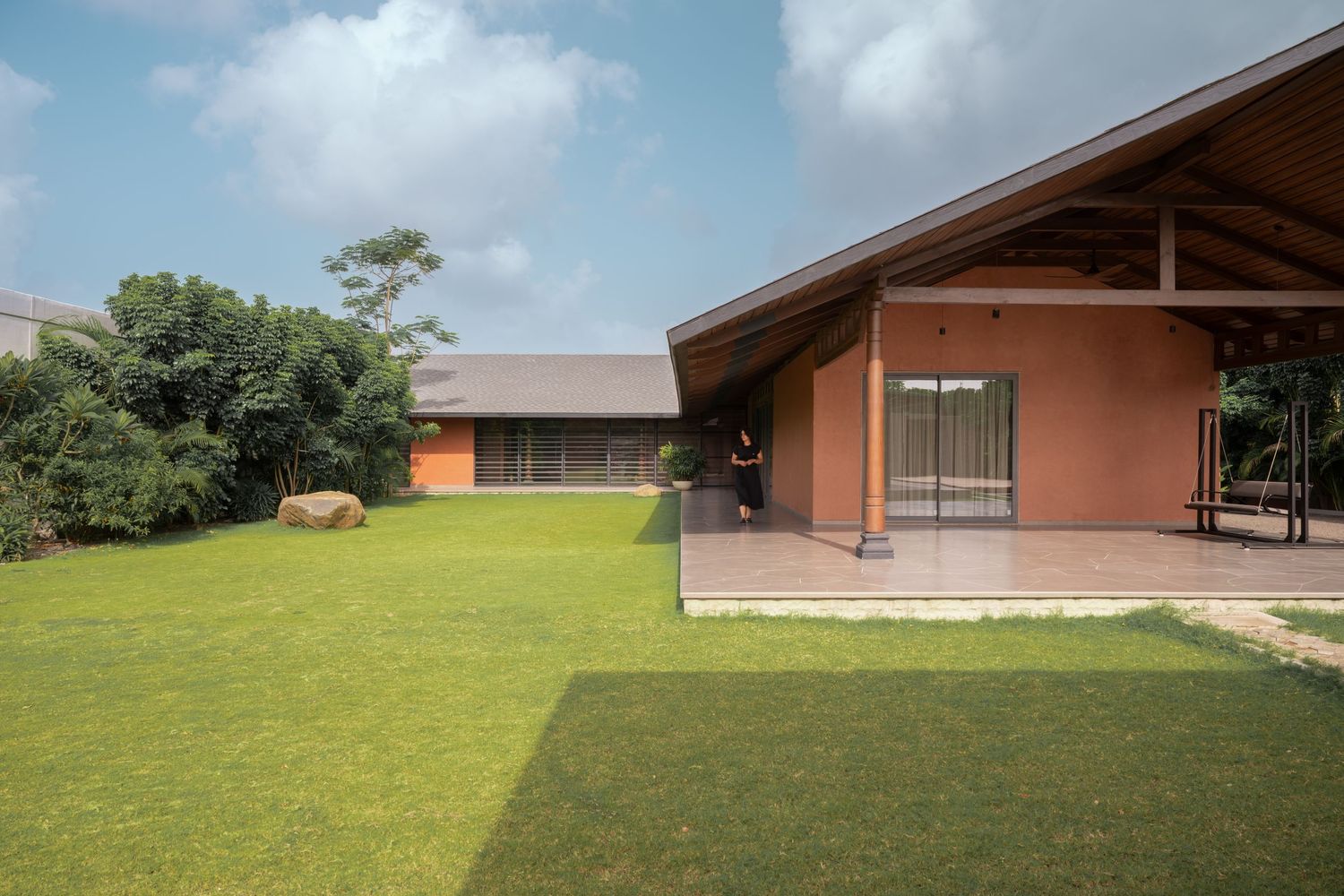 Low-profile terracotta house with exposed wooden beam roof, wide green lawn, large boulders, and tropical garden surroundings.