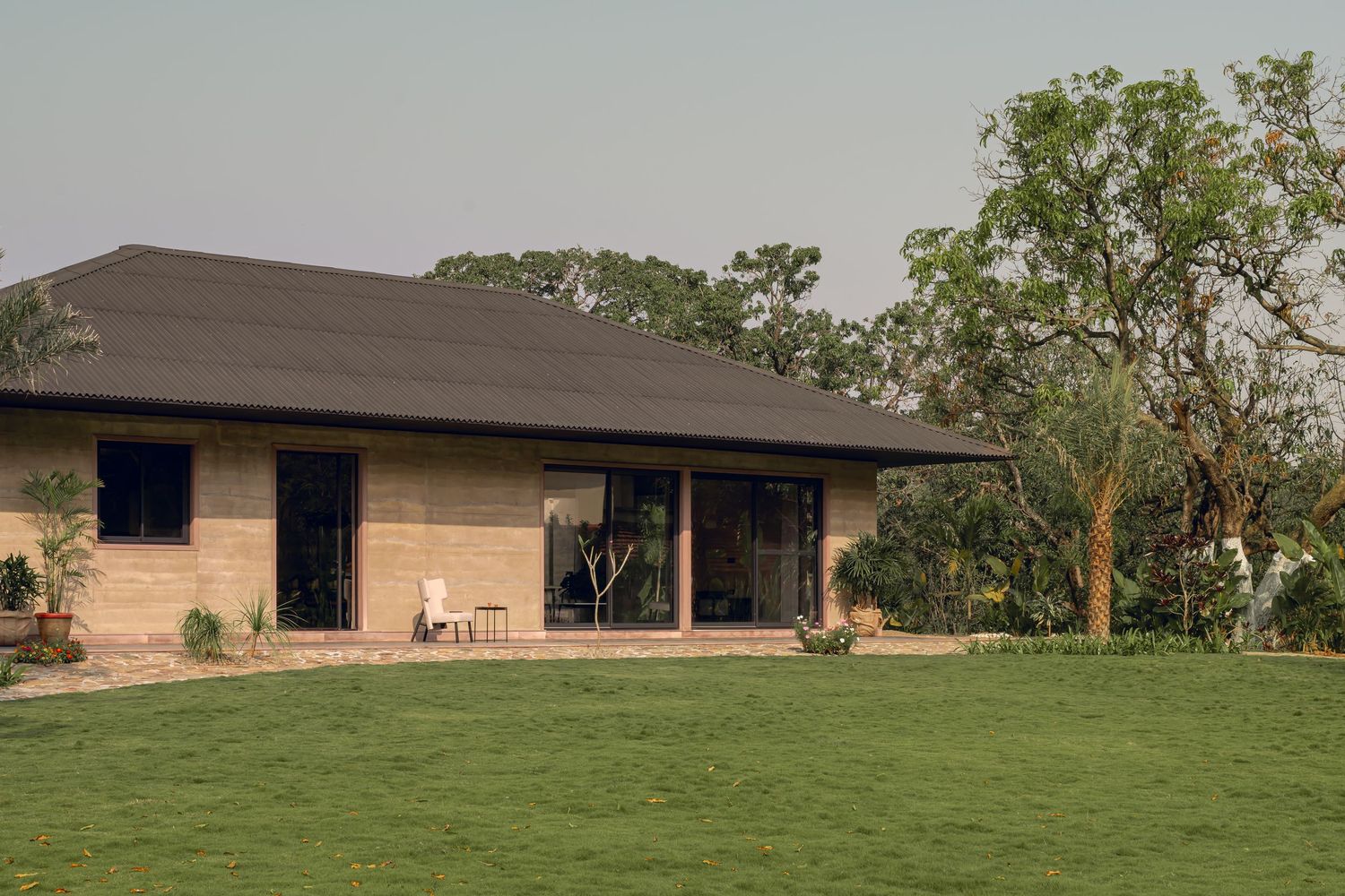 Modern single-story rammed earth home with dark pitched roof surrounded by green lawn and trees.