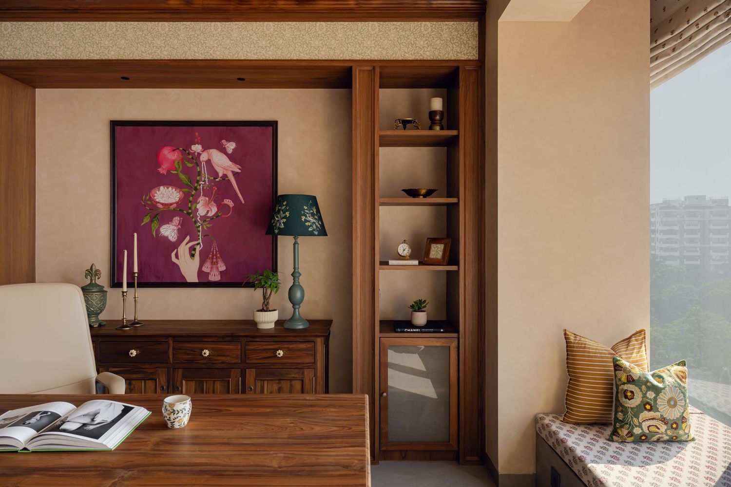 Warm wooden study nook with magenta floral artwork, teal lamp, built-in shelves, and window seat with city views.