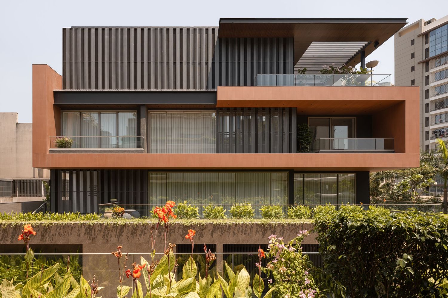 Striking modern home with terracotta and dark metal facade, horizontal balconies, and manicured green hedges.