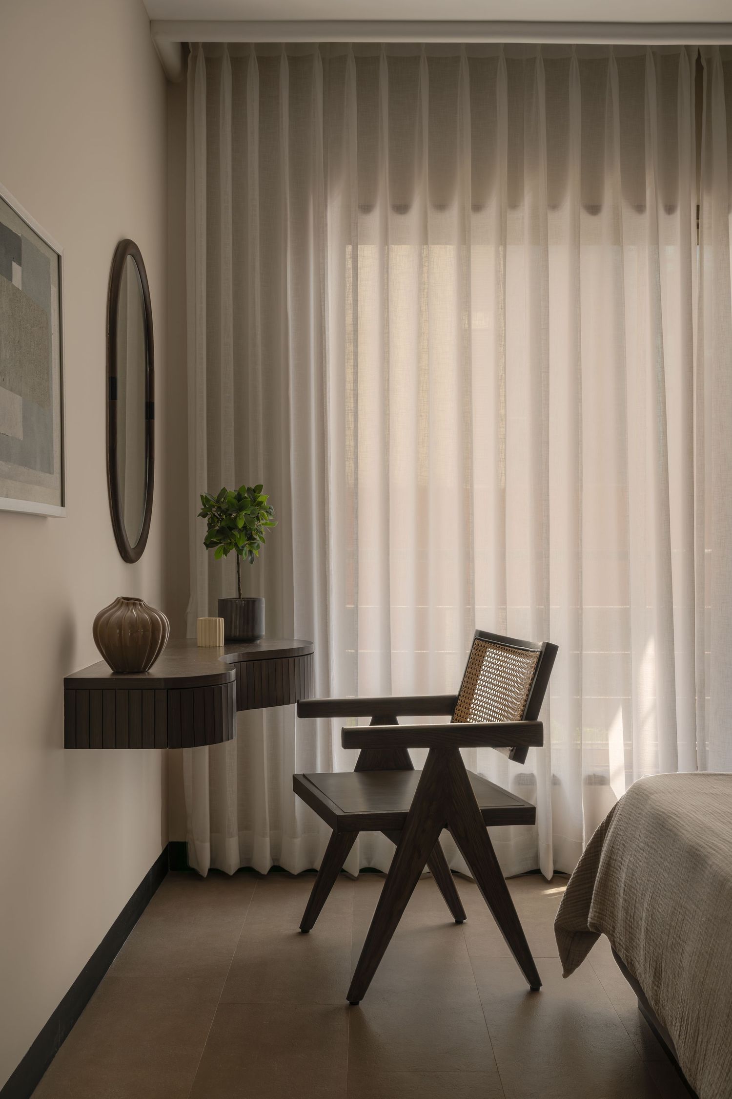 Minimalist bedroom corner with dark wood cane chair, floating console, oval mirror, and sheer curtains.