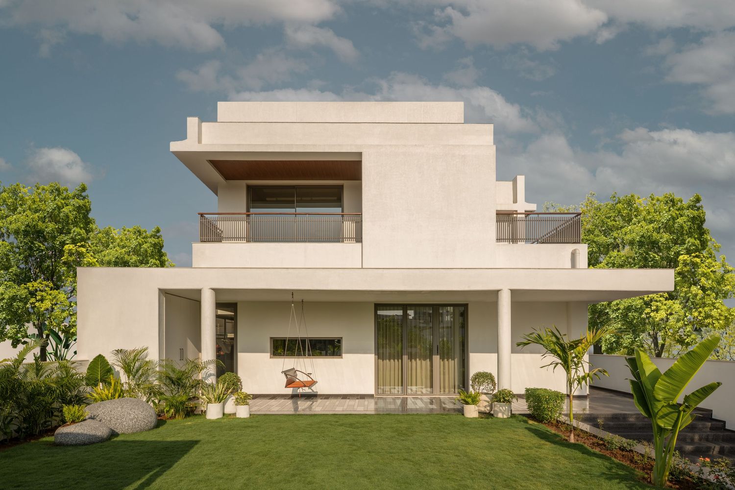 Contemporary white two-story villa with large balcony, lush green lawn, and tropical landscaping under blue sky.