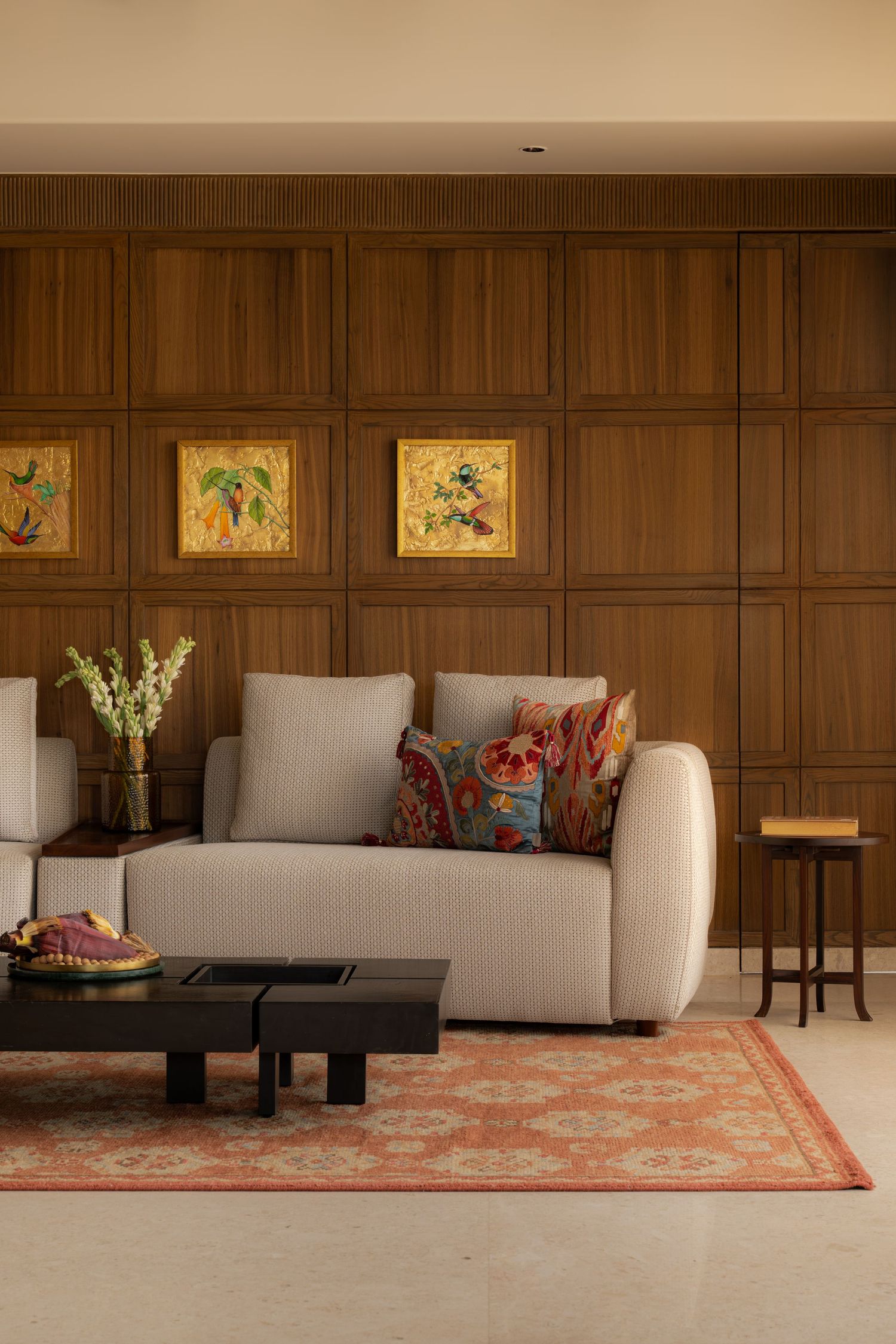 Elegant living room with beige sofa, colorful cushions, wood-paneled wall, framed artwork, and coral patterned rug.