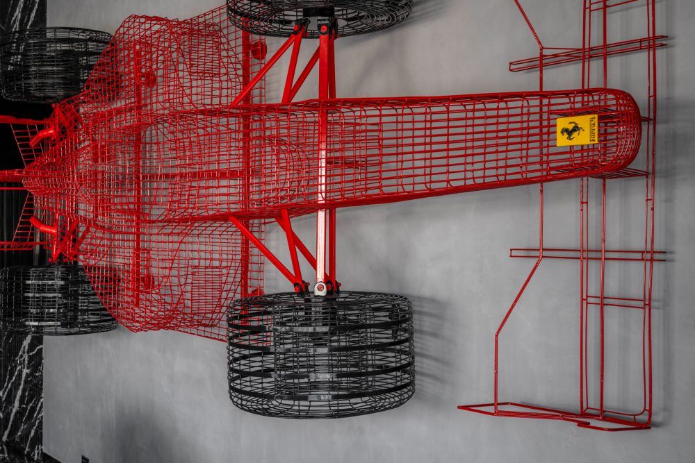 Detailed close-up of a red and black wire-frame Ferrari Formula 1 car sculpture on a gray wall.