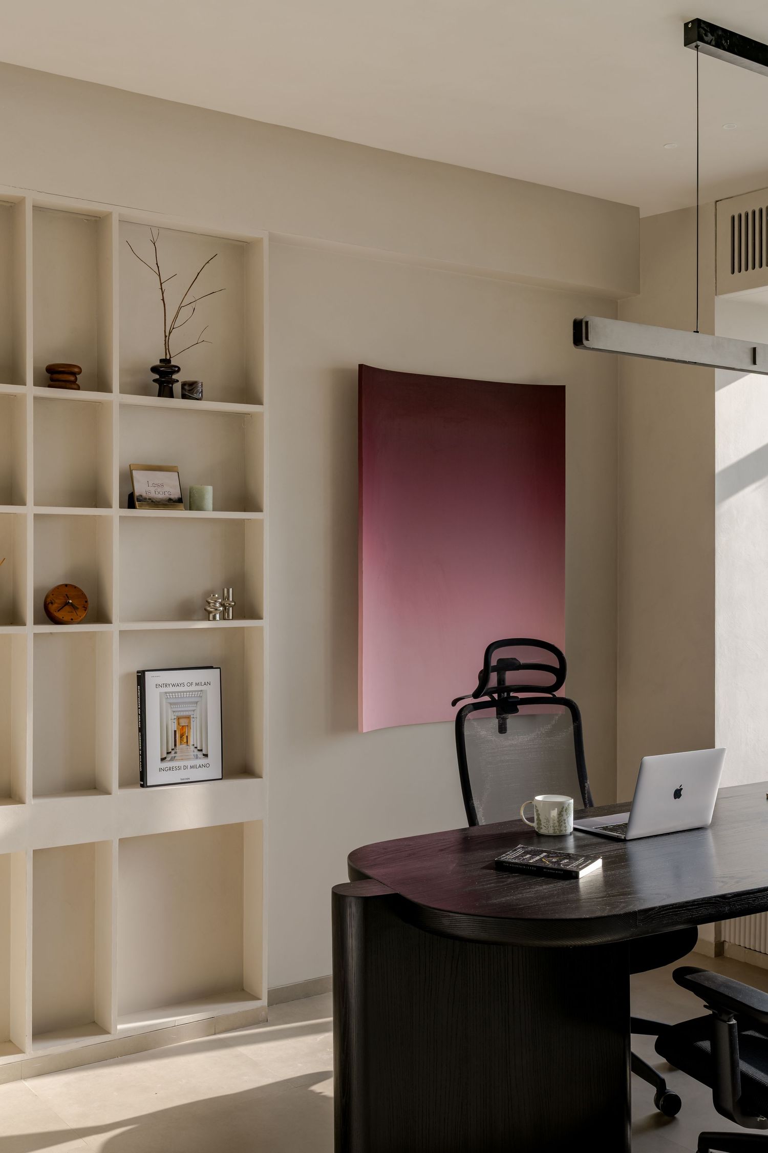 Modern home office featuring dark curved desk, laptop, ergonomic chair, cream shelving, and mauve abstract wall art.