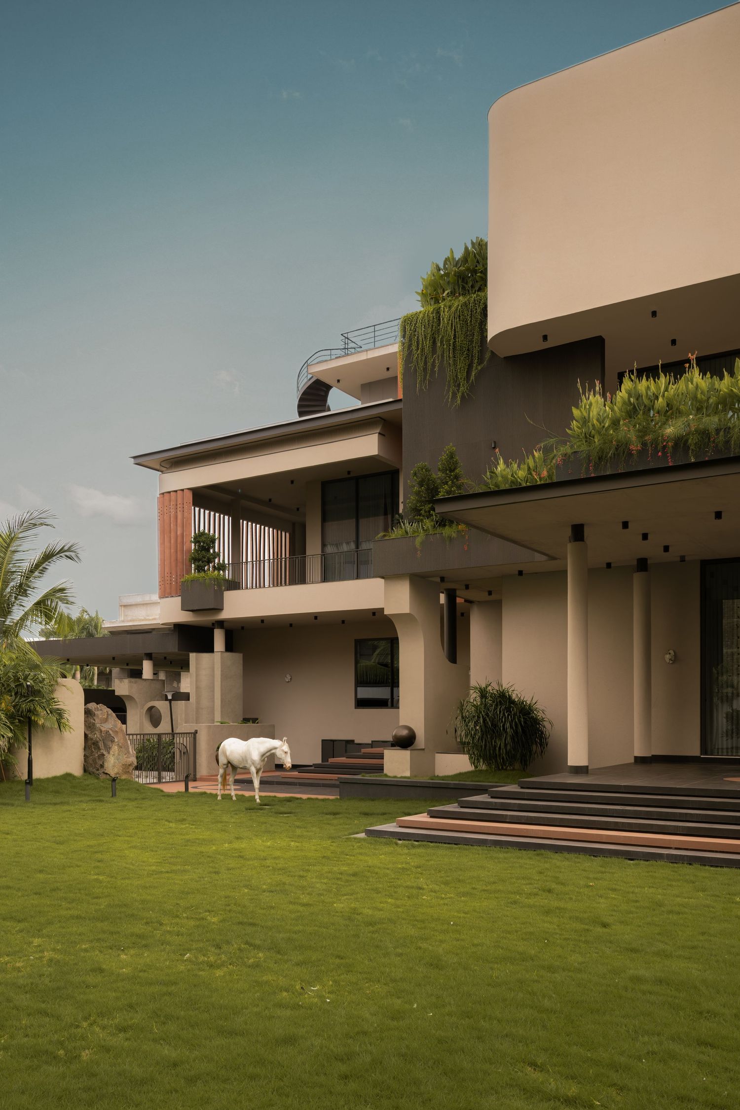 Modern two-story beige villa with rooftop garden, lush green lawn, and a white horse grazing near the entrance steps.