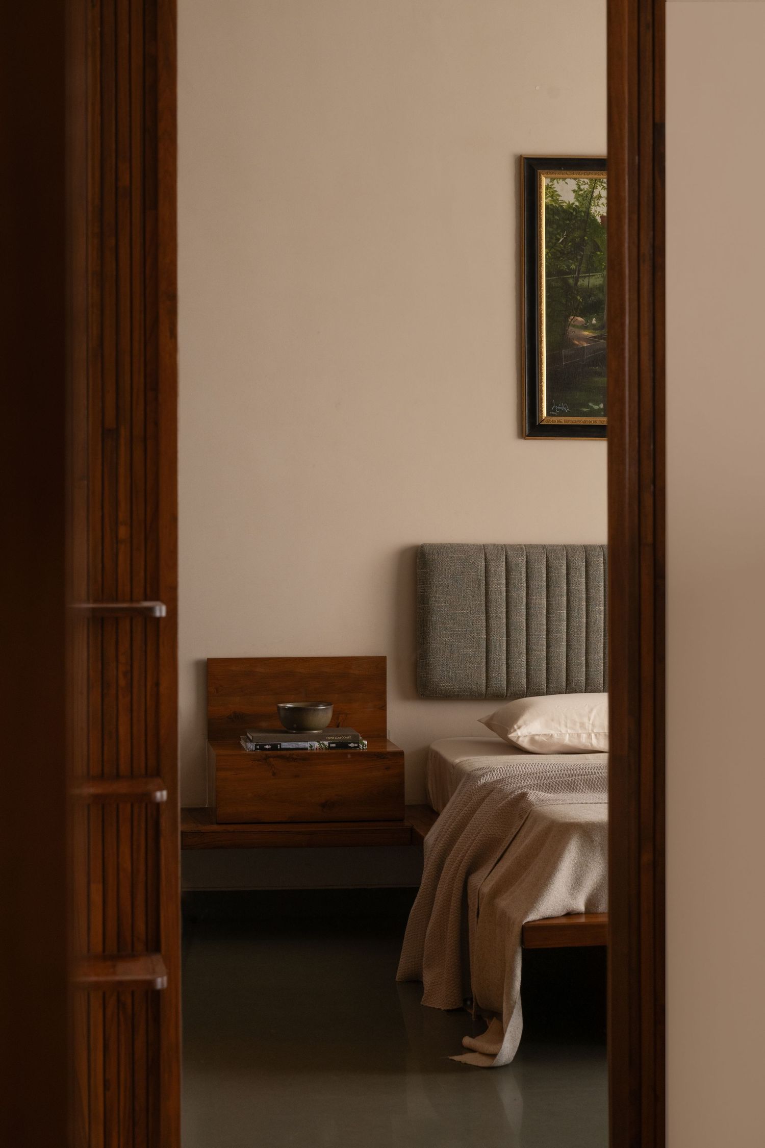Warm minimalist bedroom viewed through wooden doorframe with gray upholstered headboard and wooden nightstand.