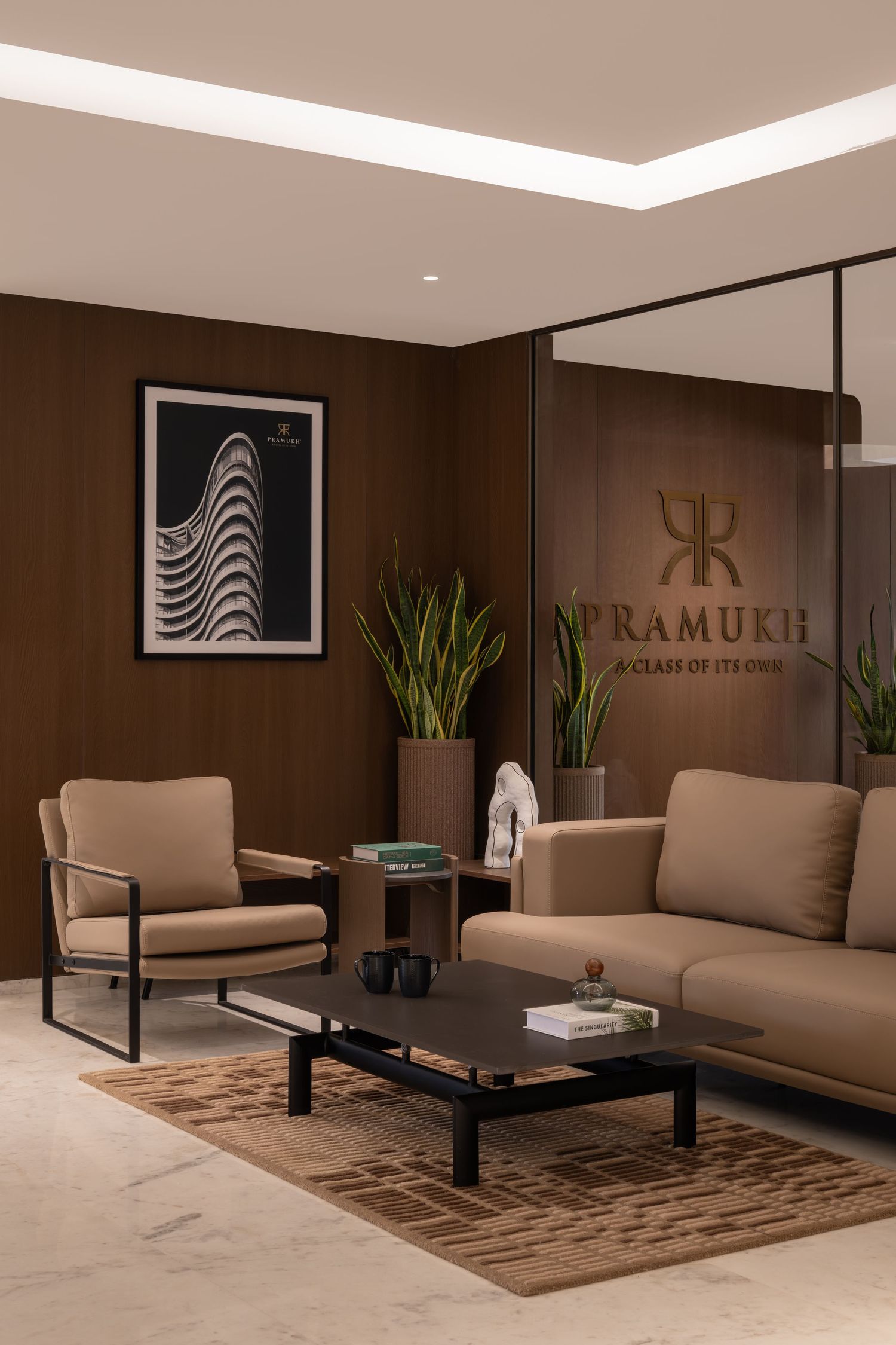 Luxury corporate reception area with Pramukh logo, beige seating, black coffee table, and dark wood accent wall.