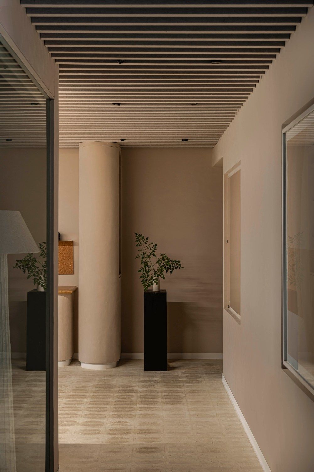 Serene beige lobby with cylindrical column, black plant pedestals, slatted ceiling, and glass partition.