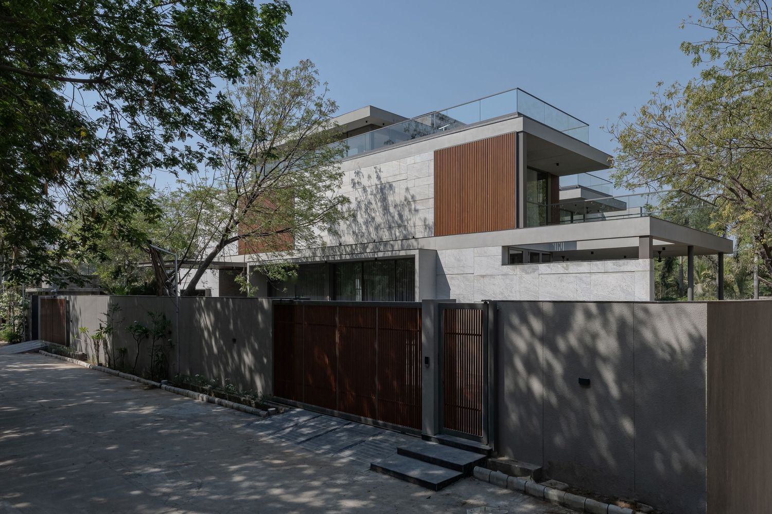 Modern two-story residence with wood and concrete facade, large gate, and mature trees.
