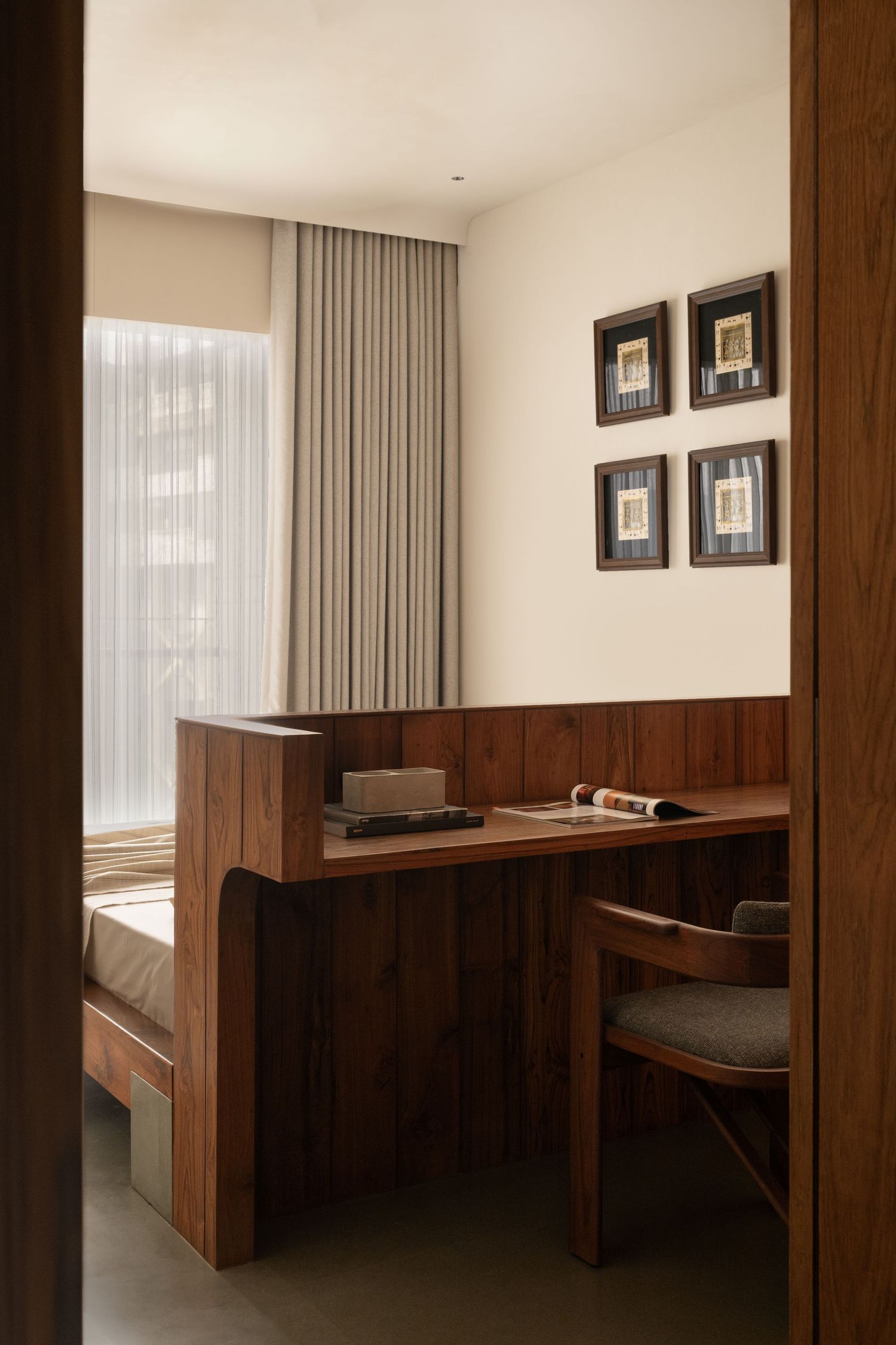 Elegant wooden home office nook with built-in desk, chair, and four framed artworks above sheer curtains.