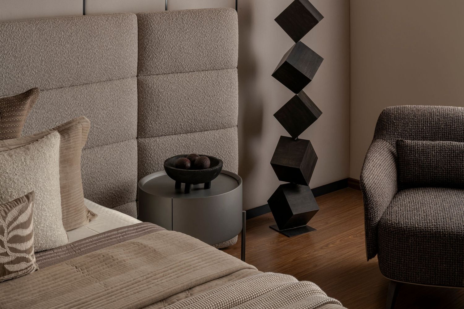 Minimalist bedroom corner featuring tufted beige headboard, dark stacked cube sculpture, round side table, and boucle armchair.
