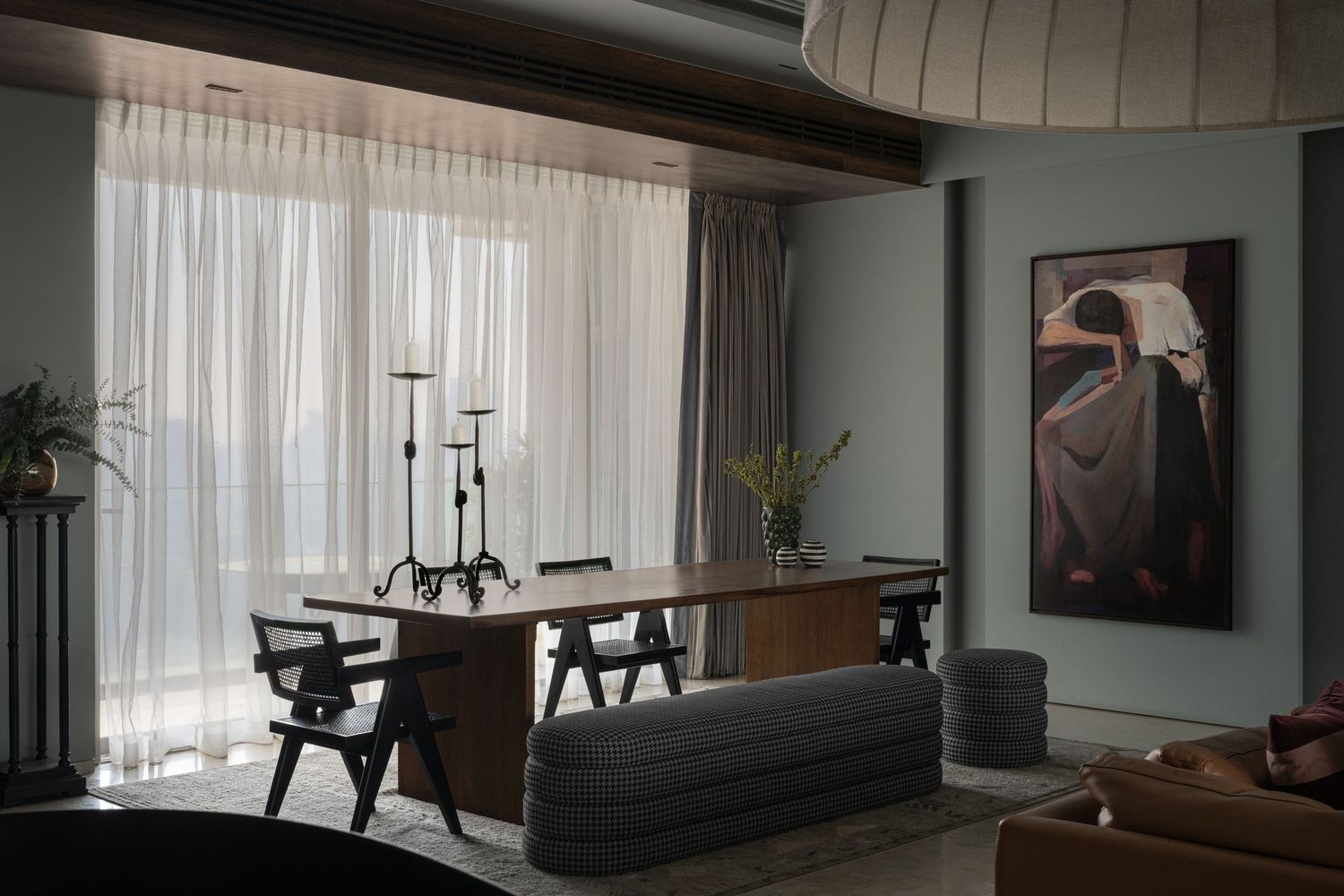 Elegant dining room with sheer curtains, wooden table, sculptural chairs, and a large figurative painting in muted tones.