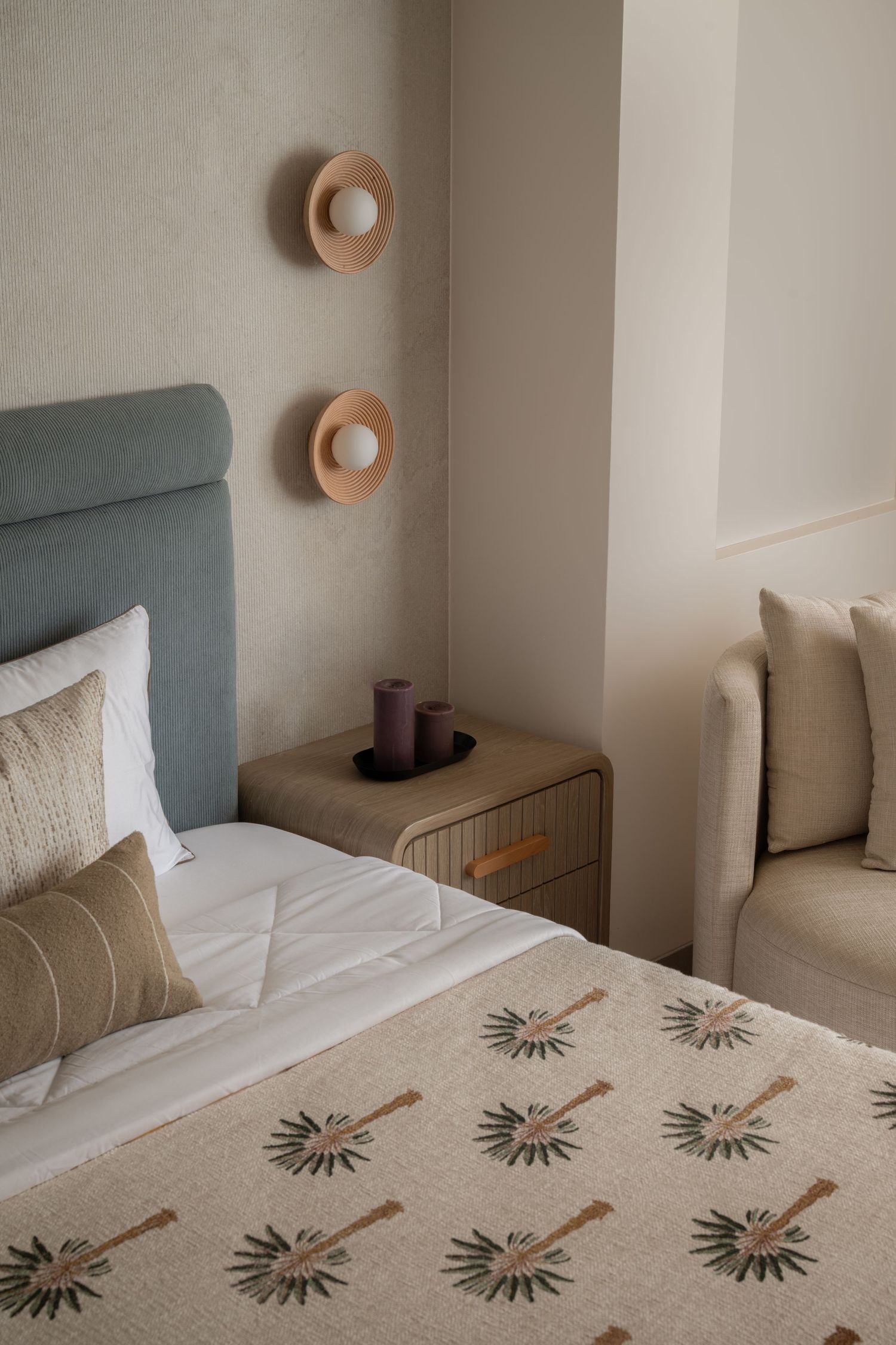 Elegant hotel bedroom with blue headboard, patterned bedspread, wooden nightstand, candles, and round wall sconces.
